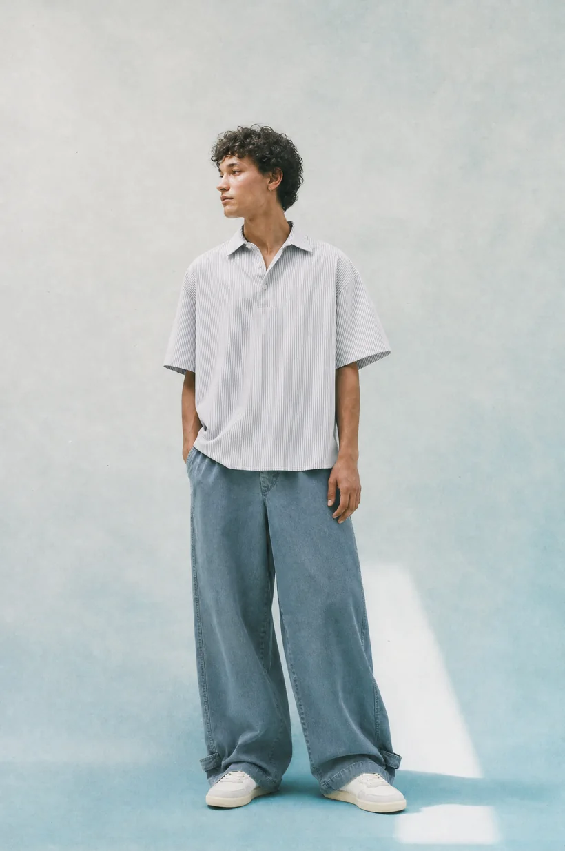Modelo de pie con polo gris y jean ancho, luz azulada de estudio