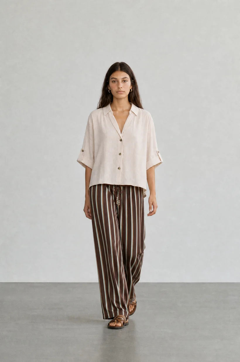 Modelo de pie con camisa crema y pantalón a rayas, fondo neutro